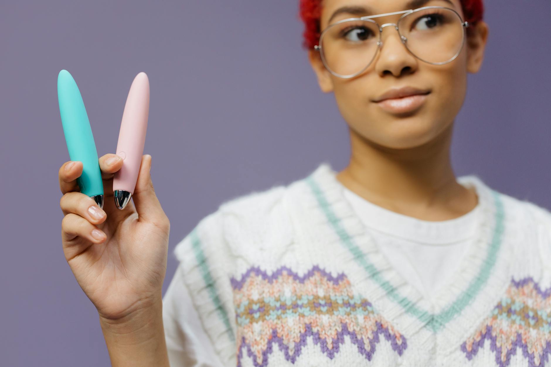 Una giovane donna con occhiali tiene due vibratori colorati contro uno sfondo viola.